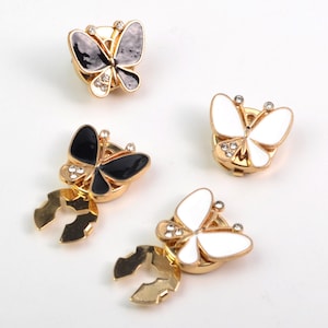 May include: Four butterfly-shaped metal fasteners with gold-tone edges. Two fasteners feature black enamel wings, and two feature white enamel wings. Each butterfly is accented with small clear rhinestones on the upper wings and a circular base.