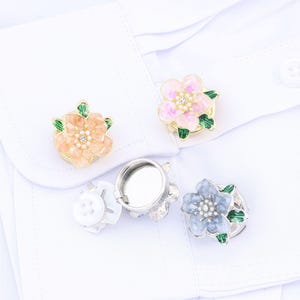Puede incluir: Tres cubrebotones en forma de flor con bordes de metal dorado, hojas de esmalte verde y detalles centrales tipo perla. Un cubrebotón es color melocotón, otro rosa claro y otro azul claro. Cada uno tiene un mecanismo de clip metálico circular.