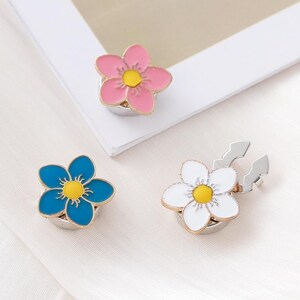 May include: Three flower-shaped magnetic clasps with enamel finishes in pink, blue, and white. Each piece features five rounded petals, a bright yellow center, and a metallic silver-toned base. The white clasp is shown with its magnetic backing visible.