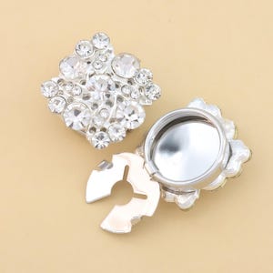 Può includere: Due bottoni metallici quadrati color argento decorati con strass trasparenti sfaccettati. Un bottone è mostrato frontalmente, evidenziando la disposizione dei cristalli, mentre l'altro è visto dal retro, rivelando un attacco metallico circolare.
