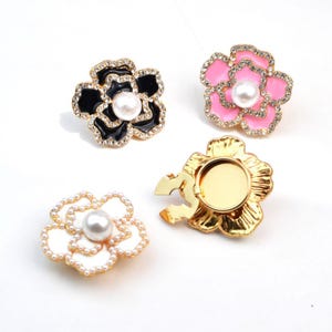 May include: Four flower-shaped phone grips in various colors. One is black with gold trim and a pearl center, another is pink with gold trim and a pearl center, a third is white with a pearl center, and the last is gold.