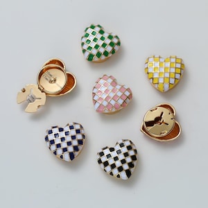 May include: A selection of heart-shaped decorative buttons in various colours, including green, pink, yellow, blue, and black, with a chequered pattern. The buttons have a gold-coloured metal border. Two buttons are open, revealing the back.