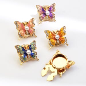 Puede incluir: Cinco broches de mariposa coloridas con metal dorado. Cada mariposa tiene una perla en el centro y está adornada con cristales transparentes. Las mariposas son en tonos rosa, naranja, amarillo, morado y azul.