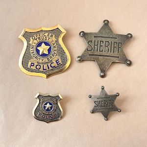 May include: Two gold-tone police badges with blue enamel centres featuring a star and the text OFFICER TRUST INTEGRITY BRAVERY POLICE, and two antique-finish metal sheriff star badges with the word SHERIFF embossed on the centre bar.