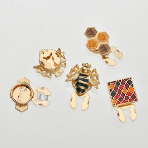 Puede incluir: Una colección de hallazgos de joyería de metal dorado. Los hallazgos incluyen una abeja, un panal, un diseño geométrico y una pieza redonda con medidas de 13 mm, 15 mm, 5 mm y 3 mm. Las piezas tienen un diseño decorativo.