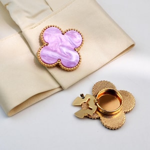 Peut inclure: Boutons de manchette en forme de trèfle avec une bordure dorée et un centre marbré violet clair. Un bouton de manchette est fixé à un poignet de chemise crème, l'autre est ouvert.