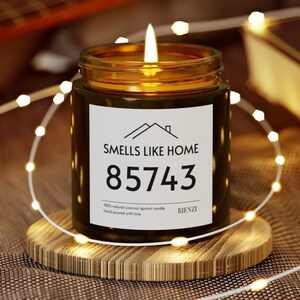 May include: A lit candle in a dark green jar with a gold rim, sitting on a wooden coaster. The label reads "SMELLS LIKE HOME 85743" with additional text. The candle is surrounded by a string of small, warm white lights.