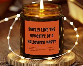May include: A lit candle in a glass jar with a gold rim, sitting on a wooden coaster. The candle has a label that reads "Smells Like The Opposite Of A Halloween Party." Decorative string lights surround the candle.