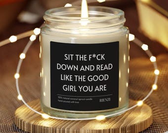 May include: A lit candle in a glass jar with a black label. The label reads "SIT THE F*CK DOWN AND READ LIKE THE GOOD GIRL YOU ARE." The candle sits on a wooden coaster, surrounded by string lights. The candle is made of 100% natural coconut apricot wax.