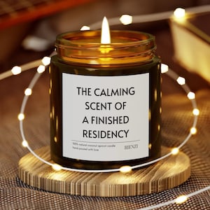 May include: A lit candle in a dark green glass jar with a gold rim, sitting on a wooden coaster. The label reads "THE CALMING SCENT OF A FINISHED RESIDENCY." A string of fairy lights encircles the candle, creating a warm, inviting ambiance.
