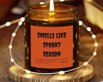 May include: A lit candle in a glass jar with a gold rim, sitting on a wooden coaster. The candle has a label that reads "Smells Like Spooky Season." The candle is surrounded by string lights.