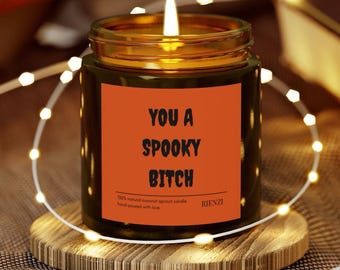 May include: A lit candle in a glass jar with a gold rim, set on a wooden coaster. The candle has an orange label with the text "YOU A SPOOKY BITCH". The candle is surrounded by a string of fairy lights. The candle is a 100% natural coconut apricot candle.