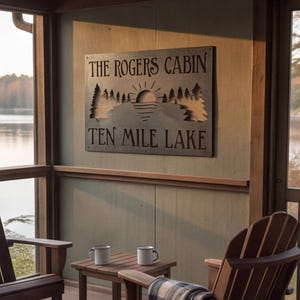 Könnte beinhalten: Ein rechteckiges Wand-Schild aus dunklem Metall mit einem lasergeschnittenen Motiv einer aufgehenden Sonne über einem See und Tannenbäumen an den Seiten. Der Text lautet THE ROGERS CABIN und TEN MILE LAKE. Das Schild hängt über einem kleinen Holztisch.