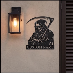 May include: A black metal wall sign featuring a hooded grim reaper figure holding a scythe, with the text CUSTOM NAME displayed in a rectangular bar at the bottom. The sign is mounted on a textured wall next to a black rectangular outdoor light fixture.