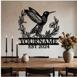 Könnte beinhalten: Ein Wandkunstobjekt aus schwarzem Metall, das einen fliegenden Kolibri zeigt, der von einem Blumenkranz umgeben ist. Unter dem Kranz befindet sich ein horizontaler Balken mit dem Text YOURNAME und EST 2024 in einer serifenlosen Schrift aus Metall.