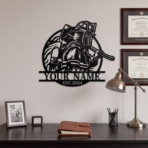 Könnte beinhalten: Ein Wandbild aus schwarzem Metall mit kreisförmigem Design, das einen Feuerwehrmann in voller Ausrüstung mit einer Axt zeigt. Unter der Figur befindet sich ein horizontaler Balken mit dem Text YOUR NAME und EST. 2024 in einer fetten, serifenlosen Schrift.