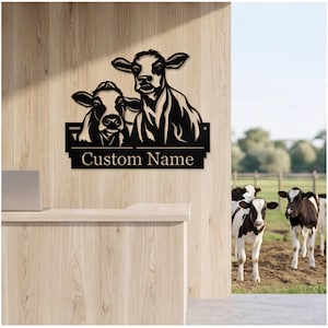 Könnte beinhalten: Ein schwarzes Metall-Wandschild mit einer Silhouetten-Illustration von zwei Kühen über einem rechteckigen Namensschild mit dem Text Custom Name in einer Serifenschrift. Das Schild ist an einer hellen Holzpaneelwand befestigt.