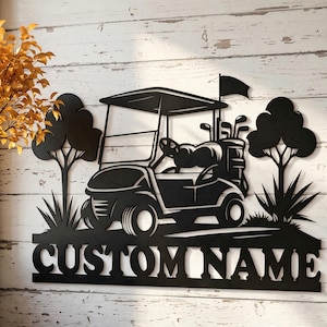 Könnte beinhalten: Schwarzes Metall-Golfwagen-Schild mit den Worten "CUSTOM NAME" am unteren Rand ausgeschnitten. Das Schild zeigt einen Golfwagen, Golfschläger, Bäume und eine Flagge. Das Schild ist auf einem weißen Holzgrund montiert.