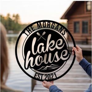 Może przedstawiać: Okrągły czarny metalowy znak z napisem THE MORGANS u góry, lake house na środku i EST 2021 na dole. Projekt zawiera grafikę przedstawiającą dwa skrzyżowane wiosła nad tekstem oraz stylizowane szczyty górskie pod tekstem.