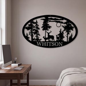 Könnte beinhalten: Ein ovales Wandbild aus schwarzem Metall mit einer lasergeschnittenen Silhouette einer Waldszene mit mehreren Hirschen, hohen Kiefern und einem kreisförmigen Mond. Der Name "WHITSON" ist in Großbuchstaben am unteren Rand des Rahmens zu sehen.