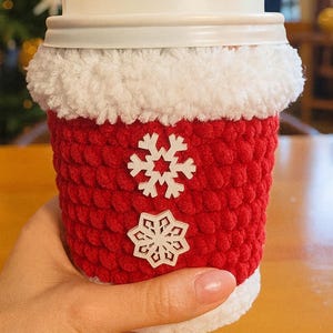 May include: A red crocheted coffee cup cozy with white snowflake appliqu&eacute;s. The cozy has a white fluffy trim at the top and bottom, and is designed to fit a standard coffee cup. A festive and decorative item.