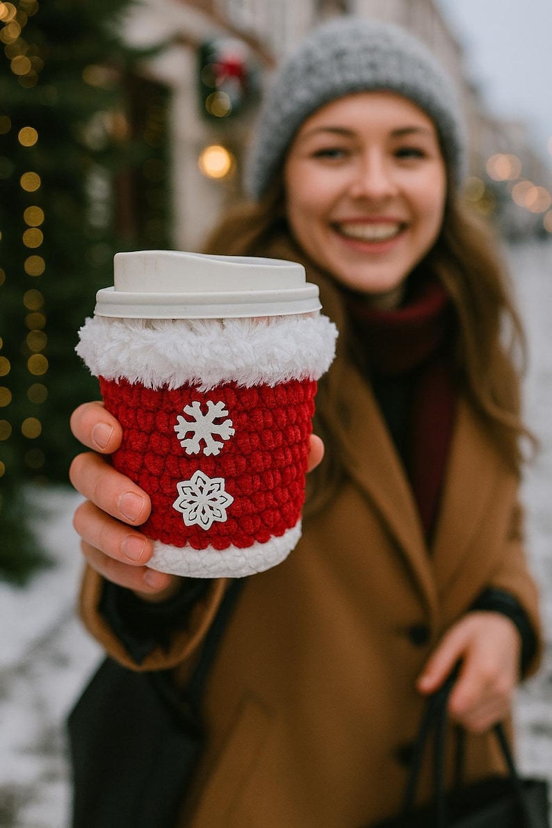 May include: A red and white crocheted coffee cup cozy with snowflake embellishments. The cozy is wrapped around a white disposable coffee cup with a lid. The cozy has a white fluffy trim around the top and bottom.