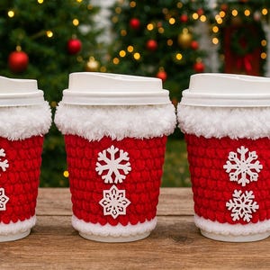May include: Three white disposable coffee cups with lids, each adorned with a red crocheted cozy. The cozies have white faux fur trim and white snowflake appliqu&eacute;s. The cups are arranged on a wooden surface with a Christmas tree background.
