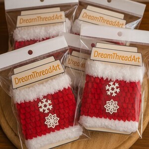 May include: Several Christmas-themed ornaments in clear plastic packaging. The ornaments are red with white trim and snowflake designs. Each package has a label that reads "DreamThreadArt".
