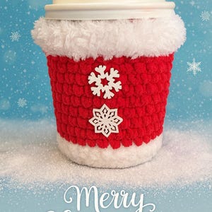 May include: A red and white crocheted coffee cup cozy with snowflake embellishments. The cozy is wrapped around a white coffee cup with a lid. The words "Merry Christmas" are written in white script on a blue background.
