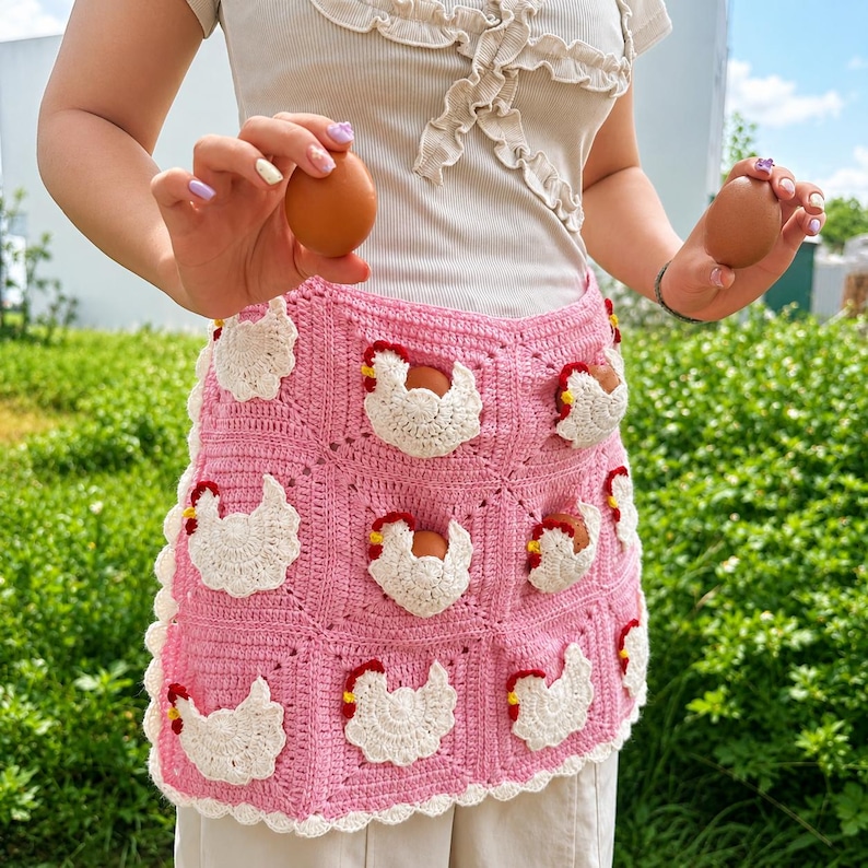 May include: A pink crochet apron featuring a grid of square panels, each decorated with a white and red chicken motif. The apron has a scalloped white border along the bottom edge and is designed with small pockets shaped like chickens.