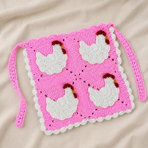 Könnte beinhalten: Eine quadratische Häkelschürze mit einer leuchtend rosa Basis, einem weißen Bogenkantenrand und zwei langen rosa Bindebändern. Vier weiße gehäkelte Hühnermotive mit roten Kämmen und gelben Schnäbeln sind gleichmäßig auf der rosa Fläche verteilt.