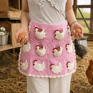 May include: A pink crochet apron featuring multiple white chicken-shaped pockets, each designed to hold a single brown egg. The apron is constructed from square panels with a scalloped white border along the bottom edge and sides.