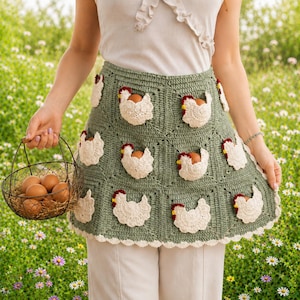 May include: A sage green crochet apron featuring multiple white chicken-shaped pockets, each containing a brown egg. The apron has a scalloped white border and is constructed from square panels. A wire basket filled with brown eggs is held nearby.