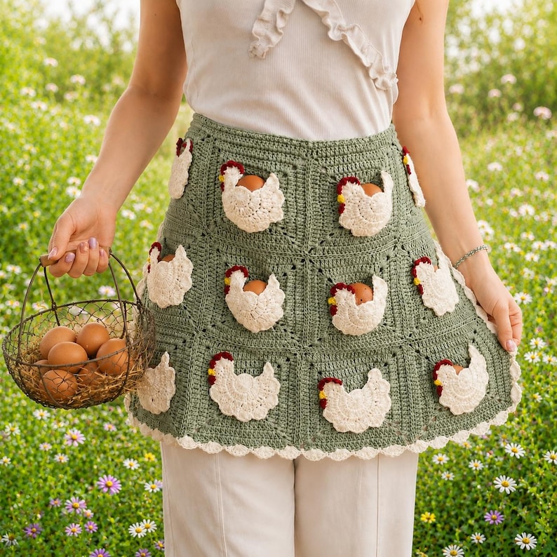 May include: A sage green crochet apron featuring multiple white chicken-shaped pockets, each containing a brown egg. The apron has a scalloped white border and is constructed from square panels. A wire basket filled with brown eggs is held nearby.