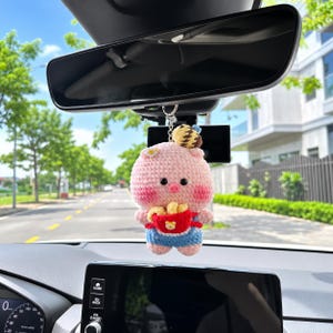 以下が含まれることがあります： バックミラーから吊り下げられた、かぎ針編みのピンクの豚のカーチャーム。豚の頭には小さな茶色のハチが乗っており、青いオーバーオールを着て、黄色いアイテムが入った赤いバスケットを持っています。黒い目、ピンクの頬、小さな白い花が特徴です。