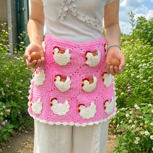 May include: A pink crochet apron featuring a grid of square panels, each adorned with a white chicken motif with a red comb. The apron includes integrated pockets designed to hold brown eggs, with a scalloped white border along the bottom edge.