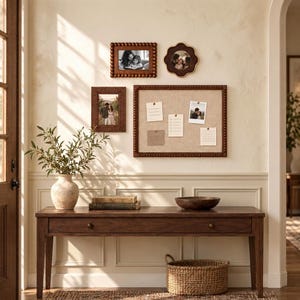 Peut inclure: Une console en bois avec un tiroir, un vase en céramique beige avec de la verdure, deux livres empilés, un bol en bois et un panier tressé. Au mur, trois cadres photo en bois et un grand tableau d'affichage en liège avec des notes.