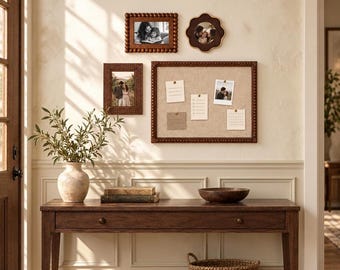May include: A wooden console table with a single drawer, a beige ceramic vase with greenery, two stacked books, a wooden bowl, and a woven basket. On the wall are three wooden picture frames and a large rectangular cork bulletin board with notes.