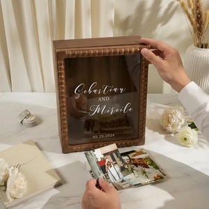 Puede incluir: Una caja de sombra de madera con frente de vidrio, con los nombres "Sebastian y Mireile" y la fecha "05.25.2024". La caja est&aacute; rodeada por un marco de cuentas. Se ven fotos y un zapato peque&ntilde;o.