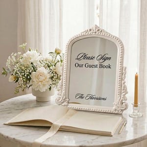 Pode incluir: Um espelho arqueado com uma moldura branca ornamentada apresentando o texto em preto Please Sign Our Guest Book e The Harrisons. O espelho é exibido ao lado de um arranjo floral branco num vaso e um livro de visitas aberto com uma caneta.