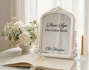 May include: An arched mirror with an ornate white frame featuring the black text Please Sign Our Guest Book and The Harrisons. The mirror is displayed next to a white floral arrangement in a vase and an open guest book with a pen on a table.