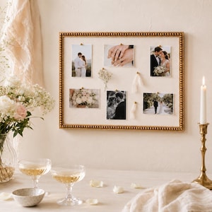 May include: A rectangular wall-mounted display board with a gold beaded frame and a cream-colored fabric backing. The board features six pinned photographs of a couple, a small sprig of dried flowers, and two short pieces of white ribbon.