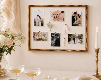 May include: A rectangular wall-mounted display board with a gold beaded frame and a cream-colored fabric backing. The board features six pinned photographs of a couple, a small sprig of dried flowers, and two short pieces of white ribbon.