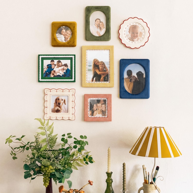 May include: A gallery wall arrangement featuring eight assorted picture frames in various shapes, colors, and textures, including velvet, scalloped, and patterned designs, displaying personal photographs of people and pets on a plain wall.