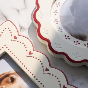 May include: Two cream-colored picture frames with scalloped edges and red trim. One frame is circular with a floral pattern, and the other is an elongated shape with a dotted heart design. Both frames contain photographs of a person and a dog.