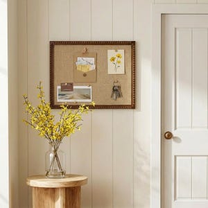May include: A rectangular bulletin board with a brown frame and a burlap surface. The board displays a small envelope, a floral print, and keys. A vase of yellow flowers sits on a wooden side table. The wall is painted white.