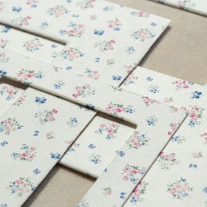 May include: Close-up of several rectangular paper cutouts with a floral pattern. The pattern features small clusters of pink, blue, and yellow flowers on a cream background. The cutouts are arranged in a layered, overlapping design.