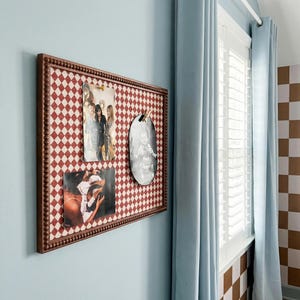 May include: A framed bulletin board with a red and white diamond pattern holds three photographs. The frame is brown with a beaded edge. The board hangs on a light blue wall near a window with light blue curtains.