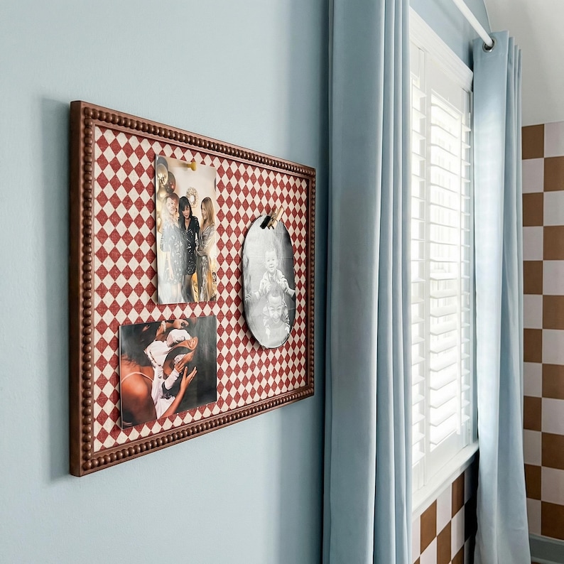 May include: A framed bulletin board with a red and white diamond pattern holds three photographs. The frame is brown with a beaded edge. The board hangs on a light blue wall near a window with light blue curtains.