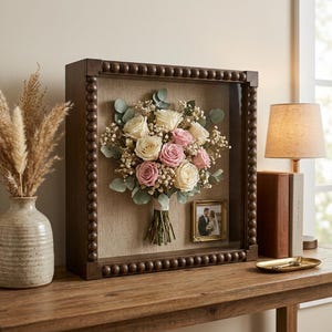 May include: A square shadow box frame with a dark brown beaded border containing a preserved bouquet of white and pink roses, baby's breath, and eucalyptus leaves. A small gold-toned picture frame holding a photograph is mounted inside.